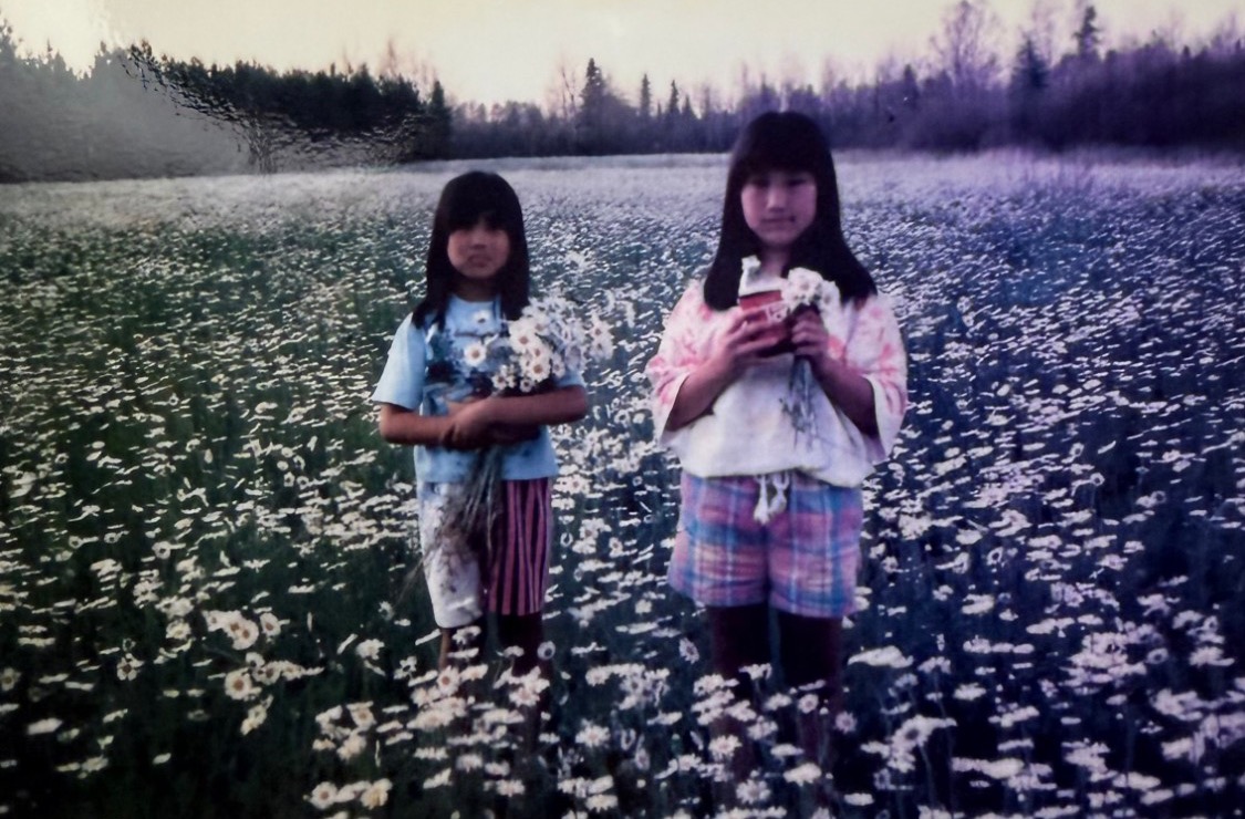 Two young girls standing in a field of daisies, each holding a bouquet
